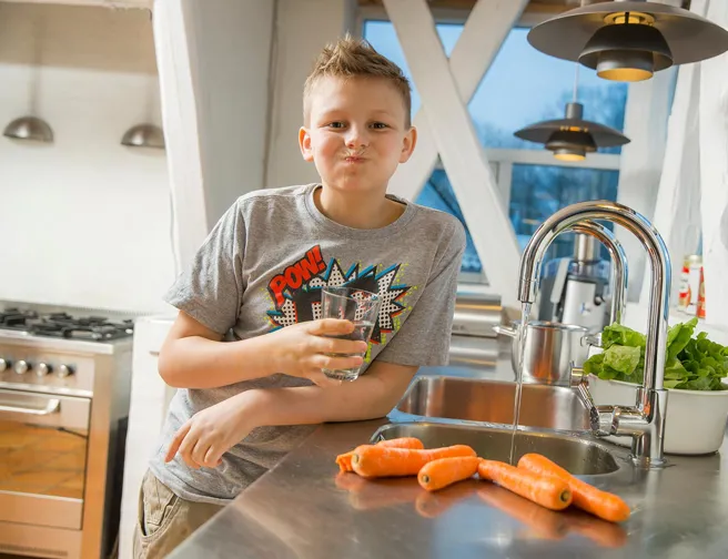 Dreng med vandglas og munden fuld af vand i et lyst køkken.