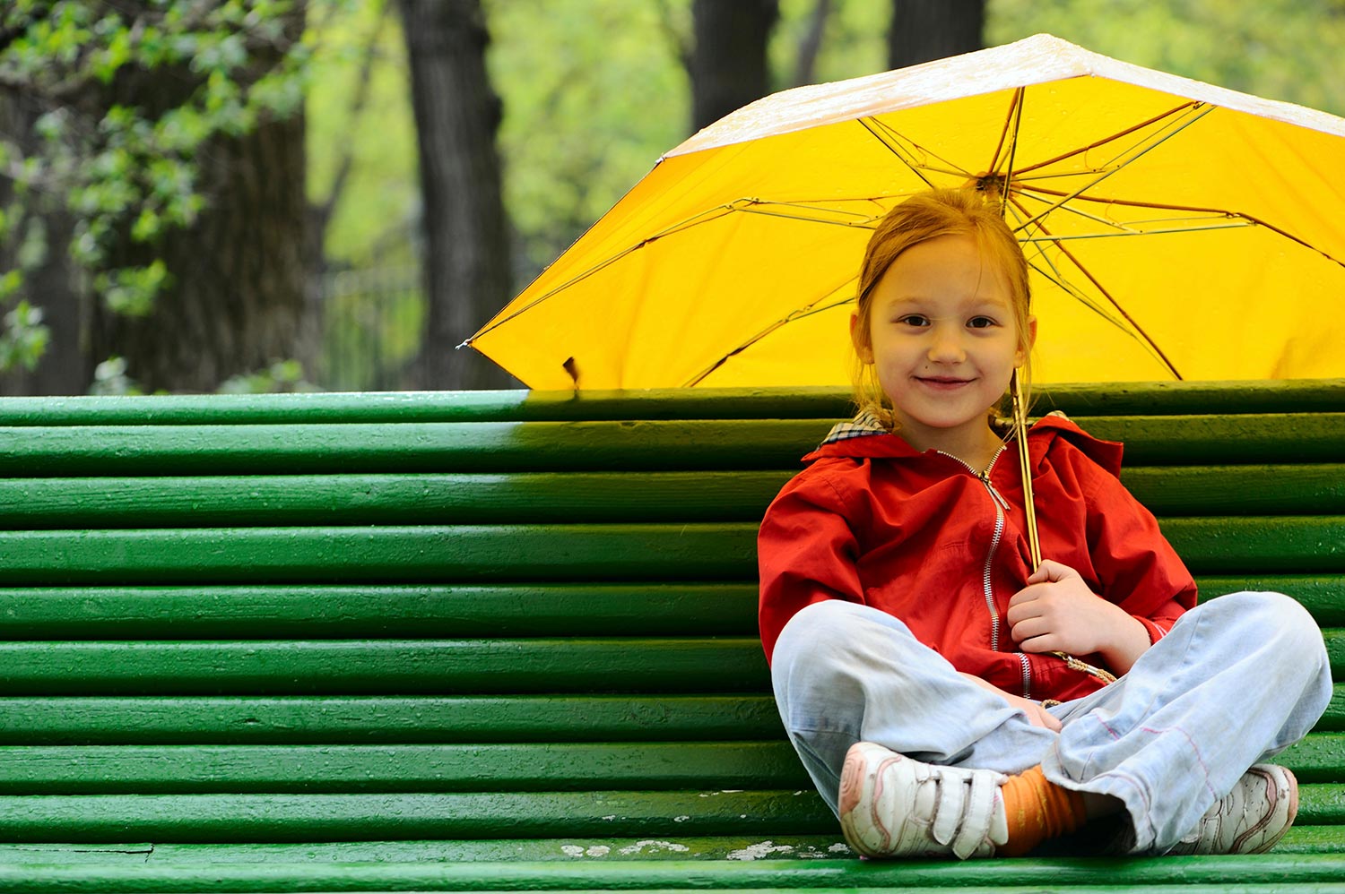 Smilende pige med gul paraply sidder på grøn bænk