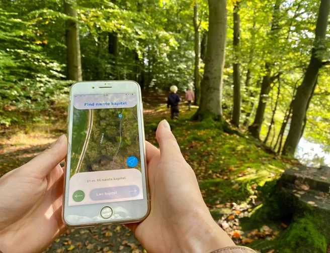 Hænder holder smartphone, der viser vej på tur i en grøn skov. To børn er løbet i forvejen.