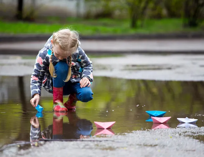 Pige leger med farvede papirsbåde i stor vandpyt